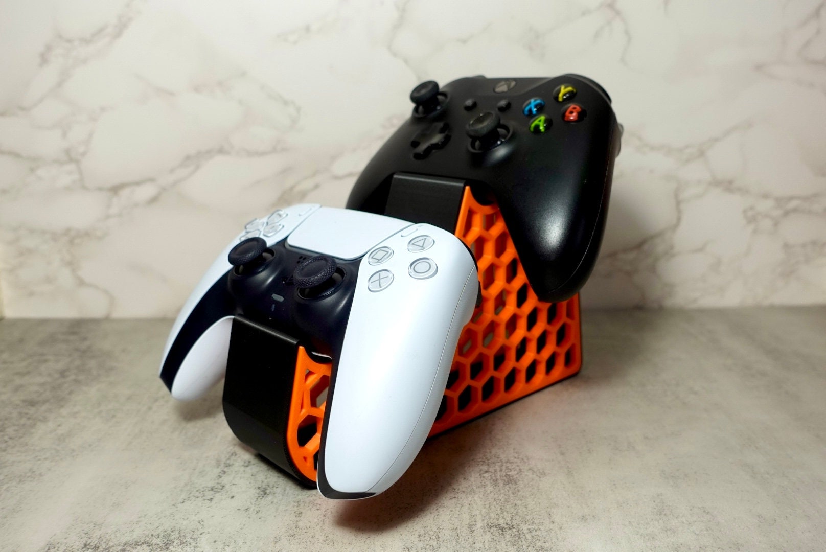 1 xbox ccontroller and 1 ps5 ccontroller placed on an orange and black holder with a honeycomb pattern on the side.