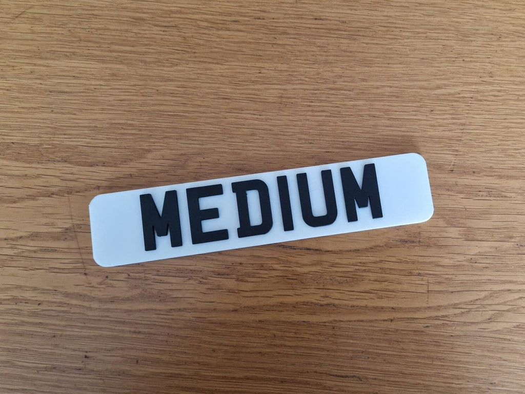 A white miniature UK car number plate with raised black letters spelling "MEDIUM" placed on a wooden surface